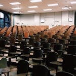 Empty university lecture hall with wooden chairs and large windows.
